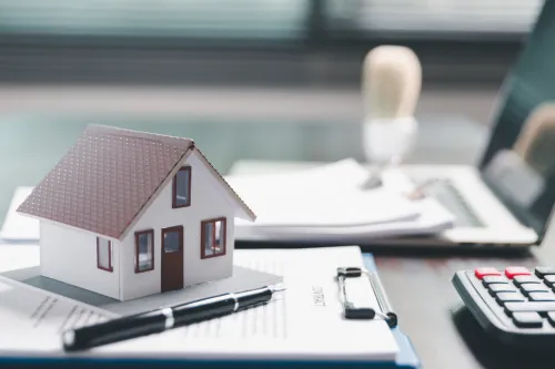 Casa em miniatura sobre documentos, com calculadora e portátil ao fundo